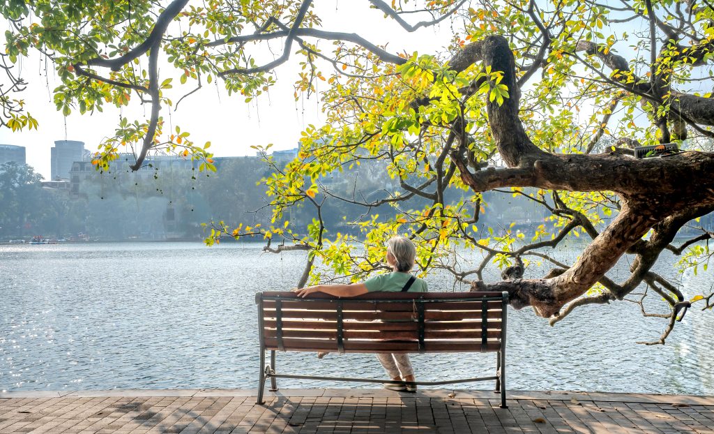 Elderly person relaxing on a bench by a tranquil lake surrounded by vibrant spring foliage.