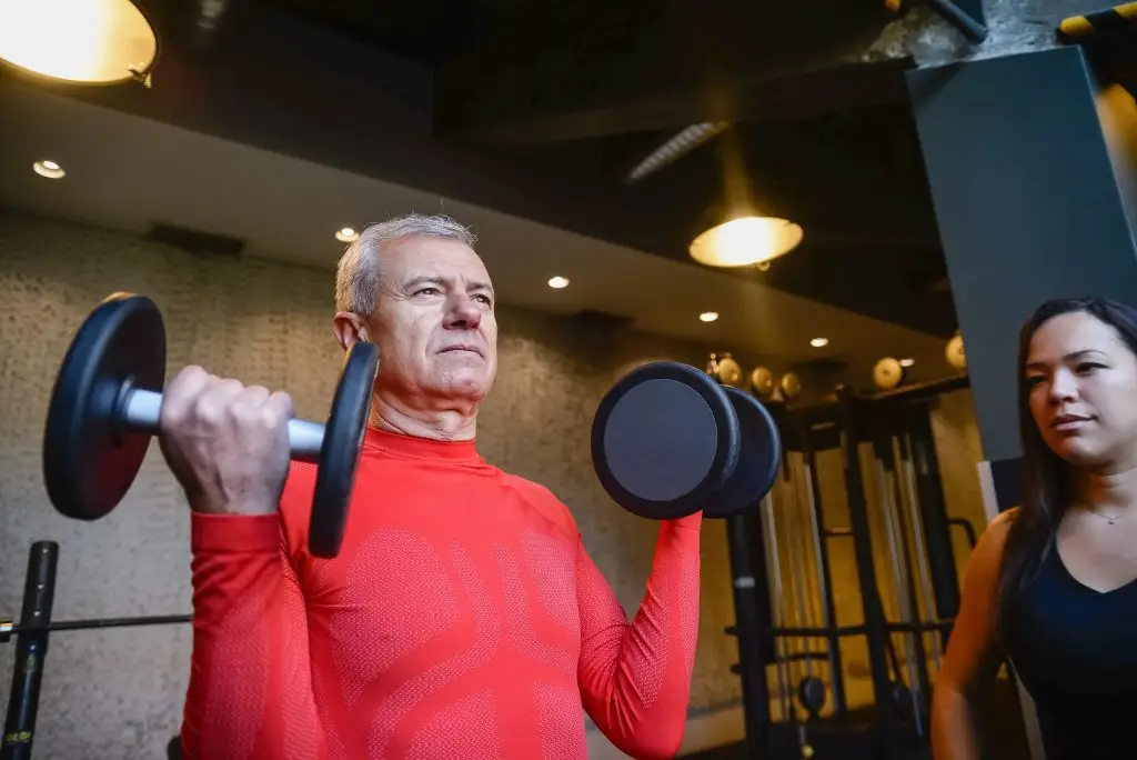 Senior man lifting weights indoors with female trainer, promoting healthy lifestyle.